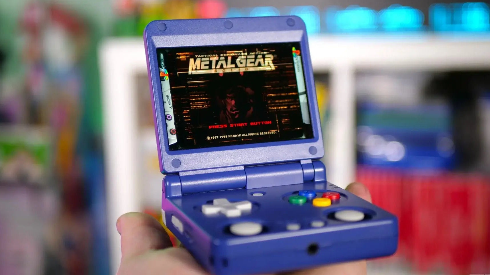 Hand holding a small blue handheld gaming console displaying 'Metal Gear', with a blurred store background.
